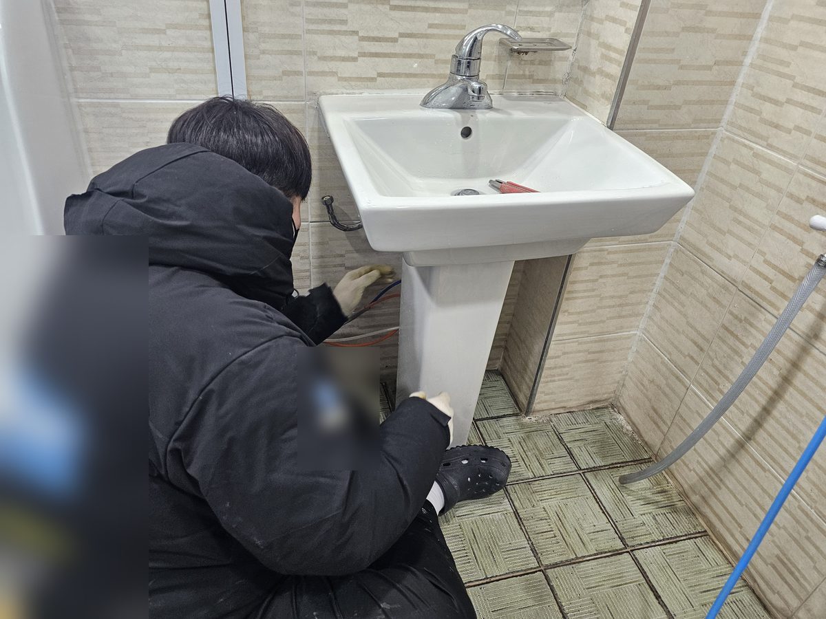 세면대 밑에 쪼그려 앉아 받침 뒤쪽 배관을 손으로 짚는 작업자