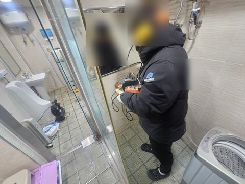 귀덮개를 쓴 작업자가 욕실 샤워부스 앞에서 탐지 장비를 손에 들고 선 모습