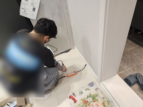 주황색 측정 장비에 이은 탐침을 벽 아래 바닥에 대고 앉은 작업자