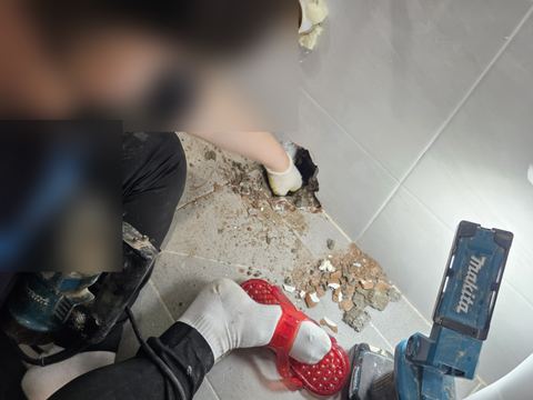 욕실 벽 모서리 하단 구멍에 손을 넣고 옆에 그라인더와 작업등을 둔 장면