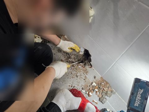 욕실 벽 하단 구멍 속 흰색 배관 둘레 모르타르를 정으로 쪼아 넓히는 모습