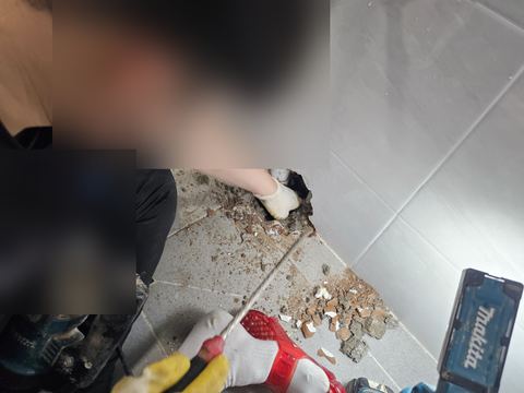 긴 정으로 욕실 벽 하단 구멍 속 흰색 배관 이음부 둘레를 긁어내는 장면