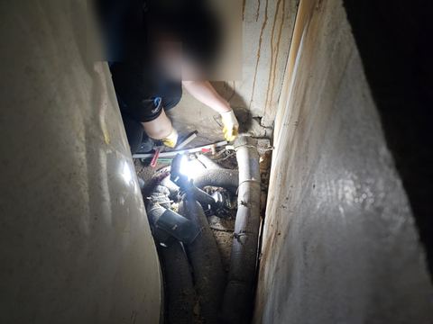 벽 사이 좁은 바닥에 놓인 보온 배관과 흰 배관, 파이프렌치 옆 벽 밑을 짚는 손