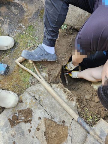 마당 바닥을 파 낸 구덩이에 손을 넣어 공구를 쓰는 작업자와 자루 긴 공구