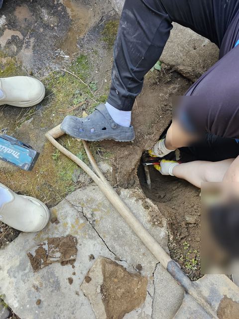 깨진 마당 콘크리트 옆 흙 구덩이에 몸을 숙여 렌치로 배관을 잡은 모습