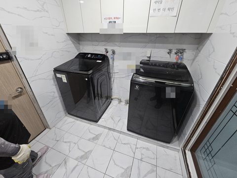 흰 타일 세탁실에 나란히 놓인 검은 세탁기 두 대와 벽의 수도꼭지