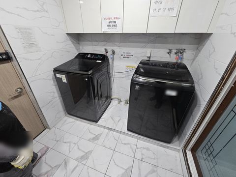 흰 대리석 무늬 벽 앞 검은 통돌이 세탁기 두 대와 급수 호스 이은 수전