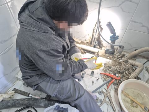 깨 낸 바닥 앞에 앉아 장갑 낀 손으로 배관 부속을 다루는 작업자