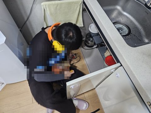 헤드폰을 쓰고 싱크대 하부장 문을 연 채 앉아 손에 든 장비를 내려다보는 작업자