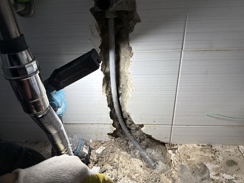 세로로 깨어 낸 벽 홈 속 회색 관과 집진기 호스, 장갑 낀 손